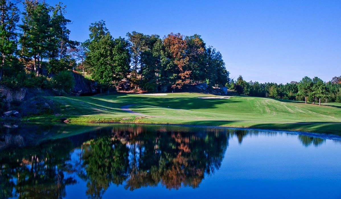 A dramatic golf hole featuring a water hazard that perfectly mirrors the autumn foliage and clear blue sky, with manicured fairways and diverse tree colors creating a stunning reflection.