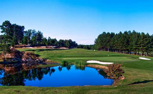 A dramatic golf hole view featuring a large pond water hazard in the foreground with lush green fairways and dense forest backdrop.