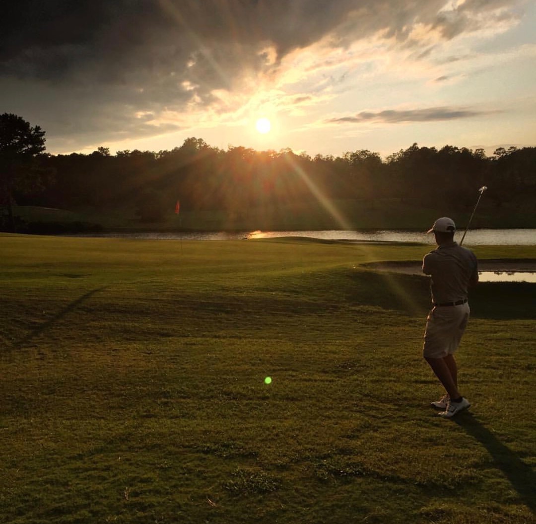 Atmospheric sunset scene with a golfer in silhouette preparing for a shot near a water hazard, creating a dramatic and inspiring golf moment with golden lighting and lens flare effects.