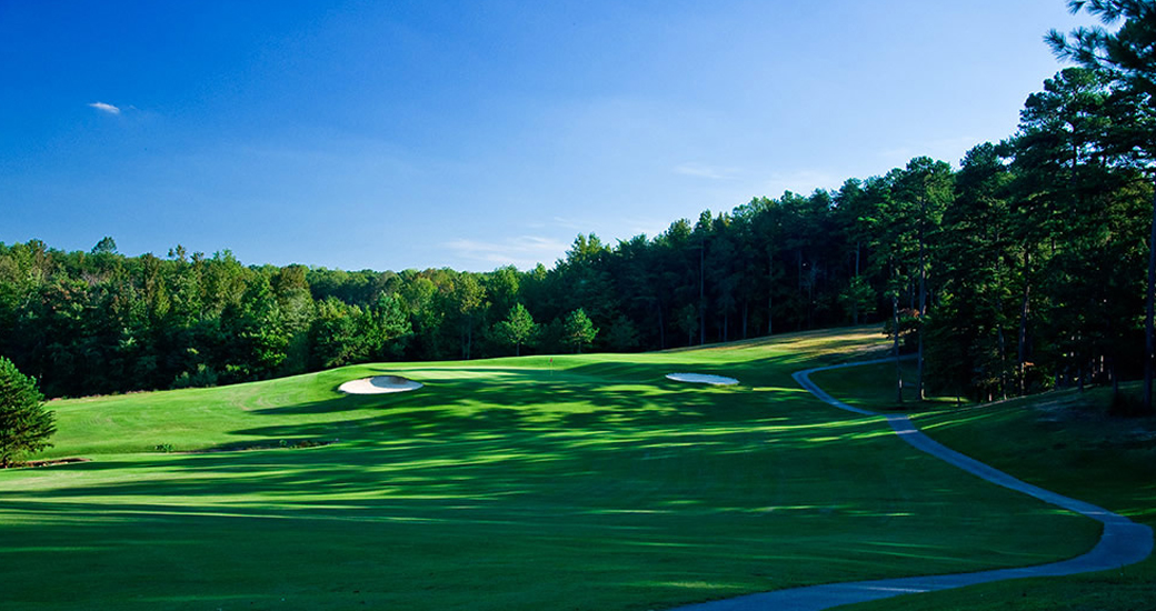 Stunning fairway view showcasing pristine green playing conditions with strategic sand bunkers, undulating terrain, and dense forest backdrop under a bright blue sky.