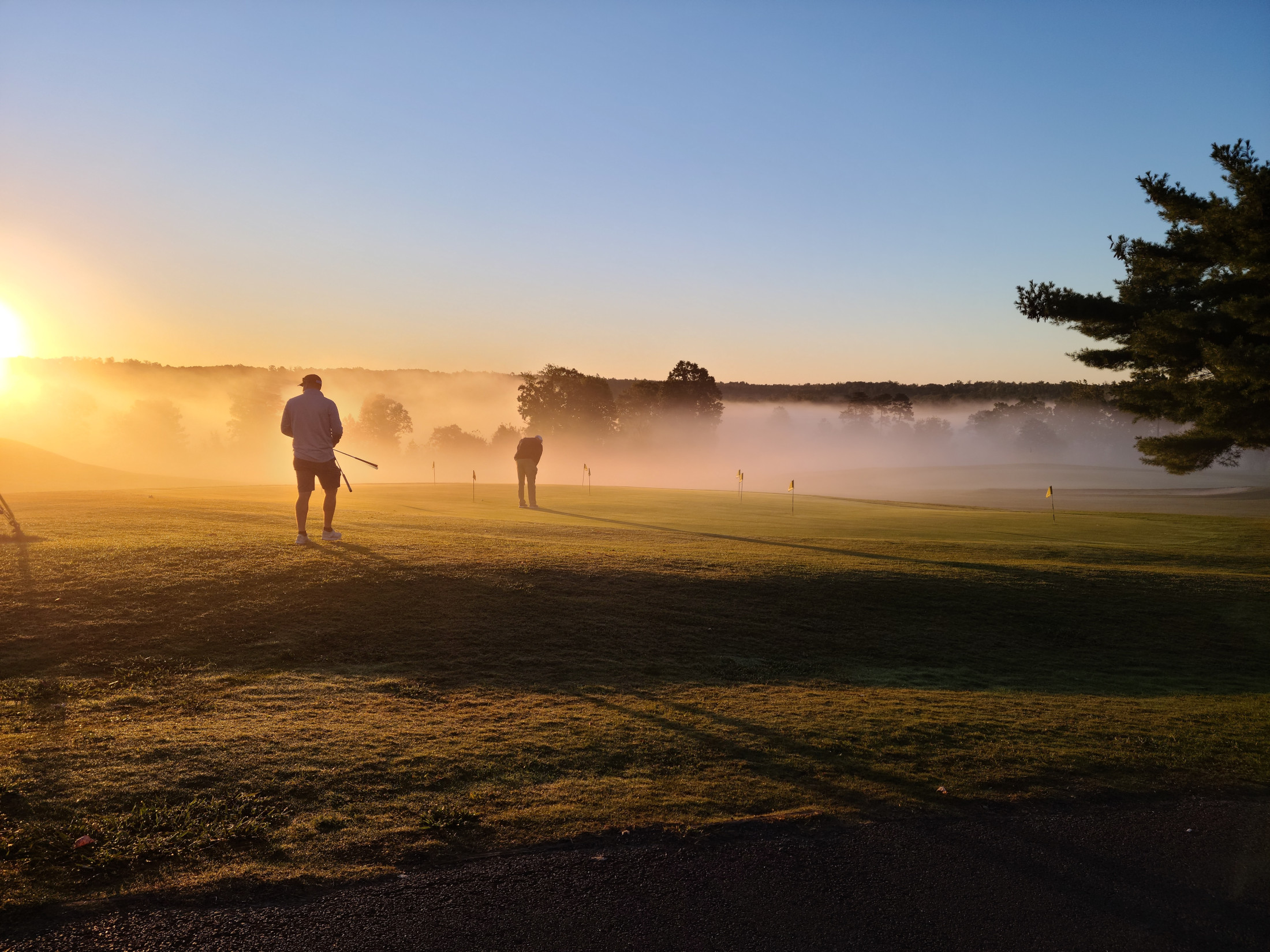 A dramatic early morning scene at Dogwood Hills showing golfers silhouetted against a golden sunrise with atmospheric mist rising from the course.