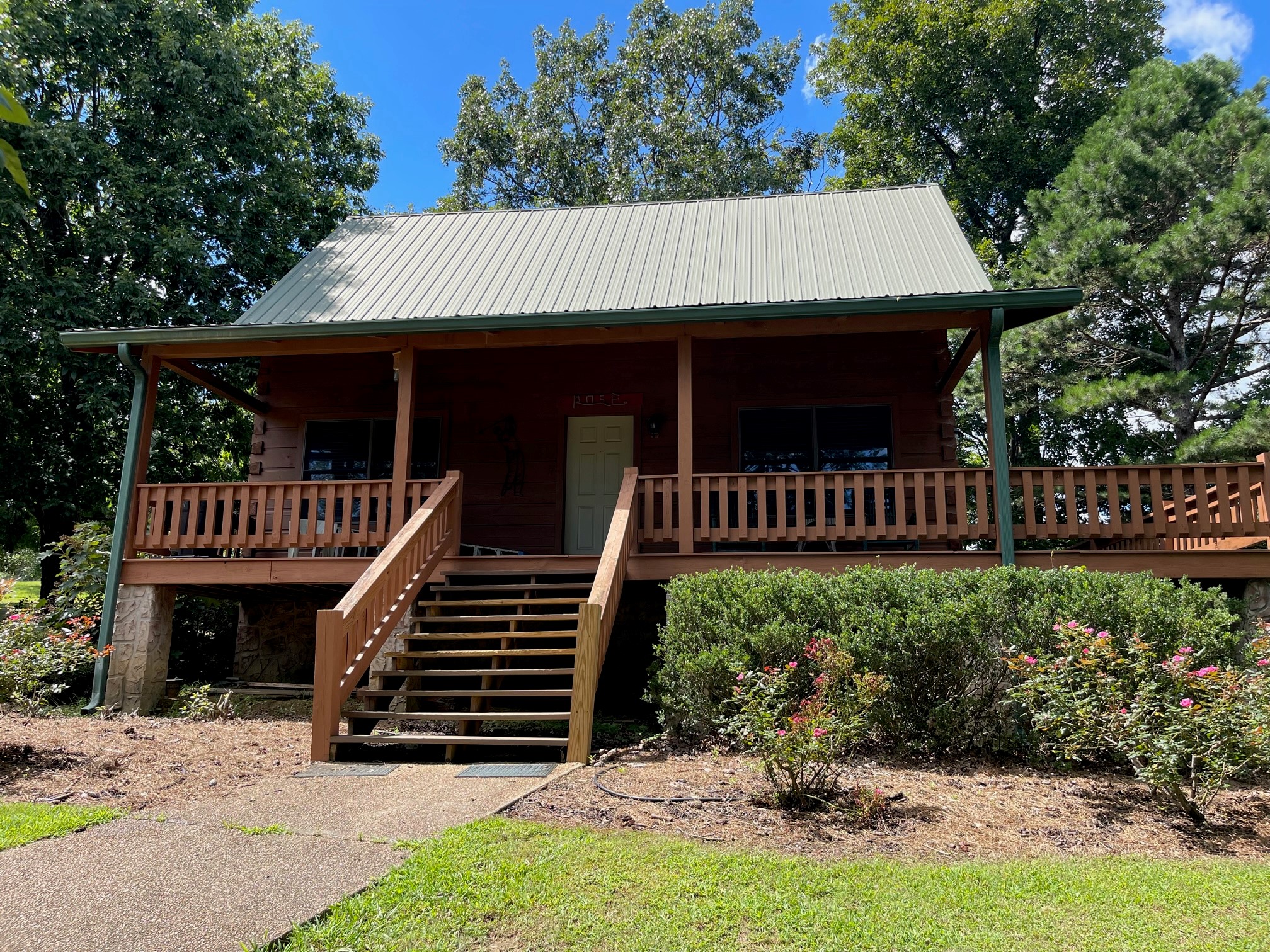 Log cabin with green metal roof, covered front porch with wooden railings and stairs, stone foundation, surrounded by landscaped gardens and mature trees.
