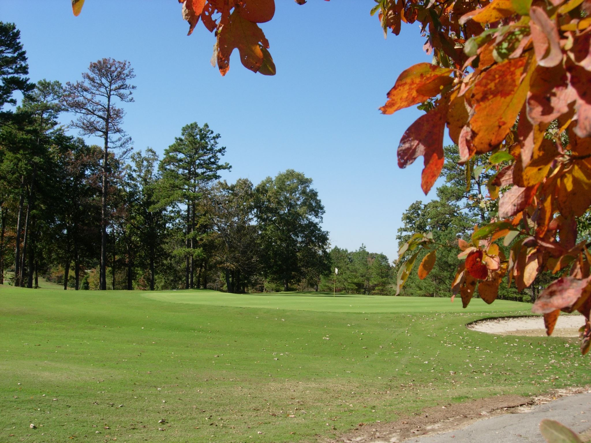 An autumn golf course view showing a well-maintained fairway with vibrant orange and red fall foliage framing the scene and mature trees in the background.