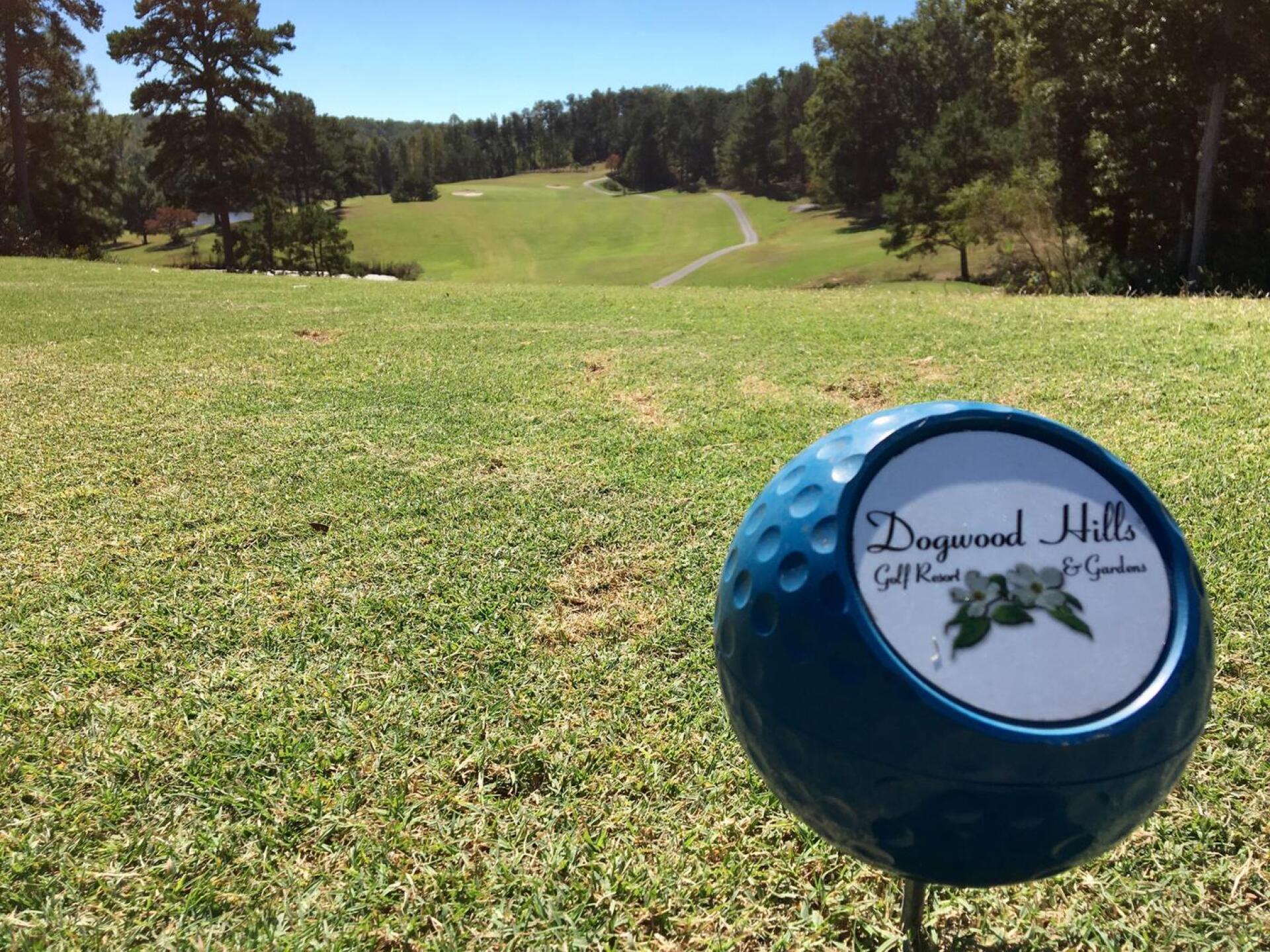 Golf course view showing rolling fairways lined with mature trees, a cart path winding through the landscape, and a branded blue golf ball in the foreground displaying the Dogwood Hills logo.