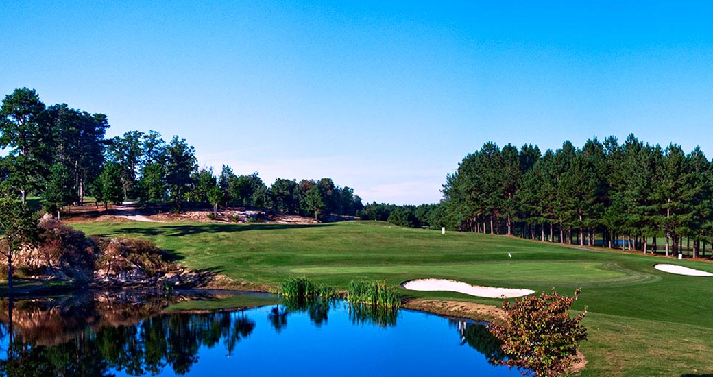 A challenging golf hole at Dogwood Hills showcasing a water hazard along the fairway with white sand bunkers, lush green turf, and dense tree coverage.