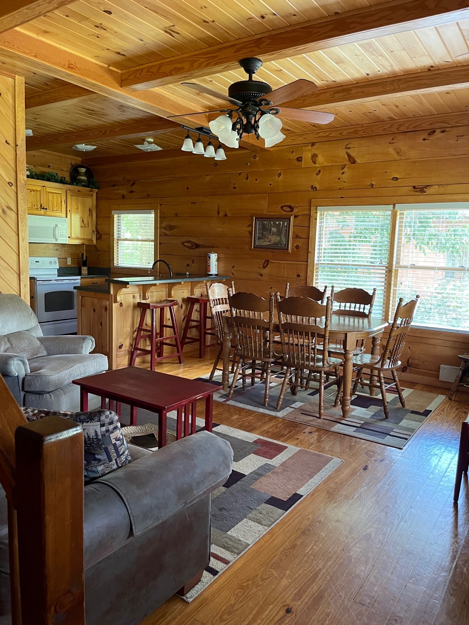 Open-concept cabin interior featuring a kitchen with wood cabinets, dining area with traditional chairs, living space with gray seating, and ceiling fan with wood beam ceiling.