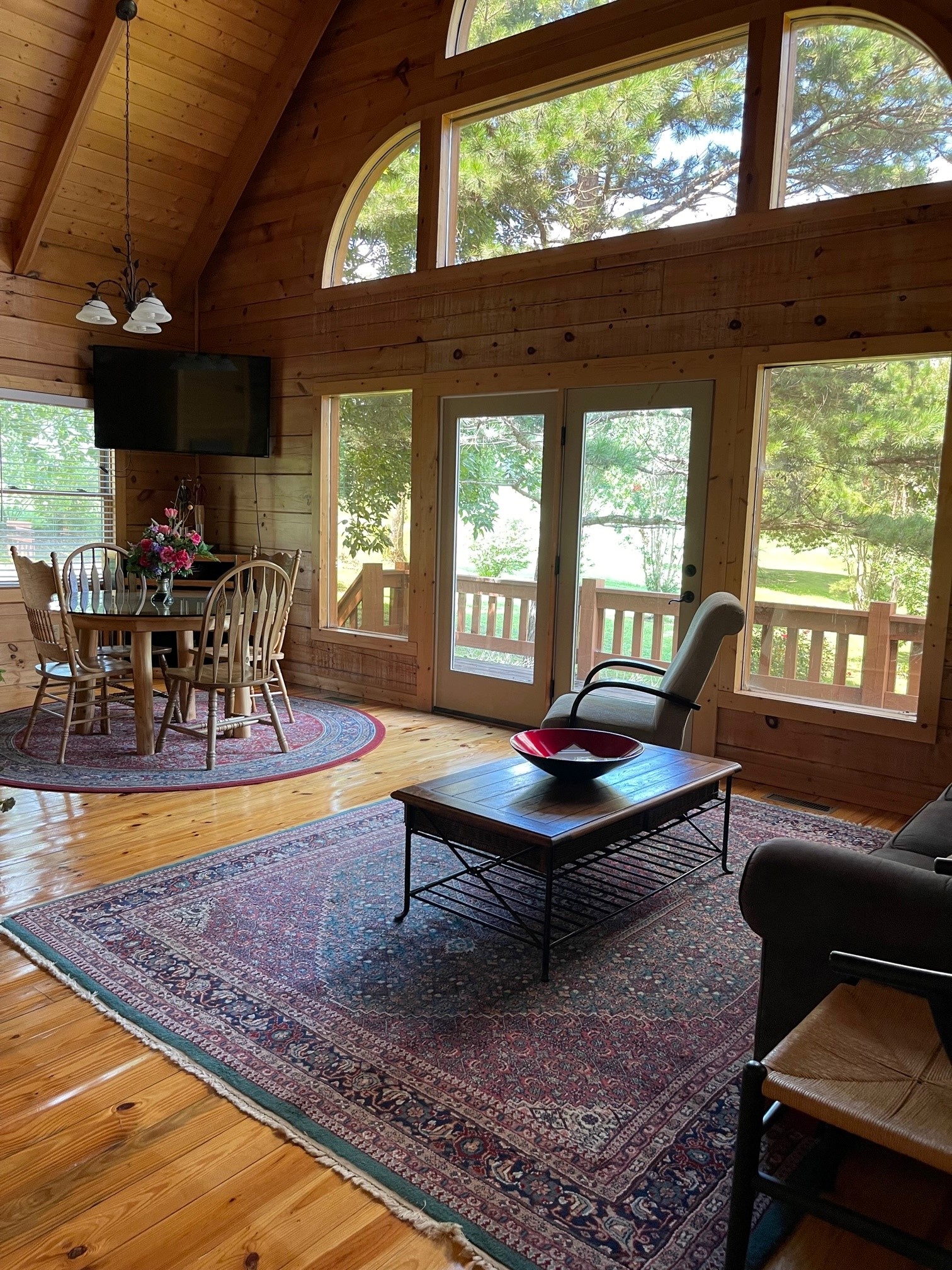 A rustic cabin living room with exposed wood beams, large arched windows overlooking trees, traditional furniture including Windsor chairs and patterned rugs, creating a cozy lodge atmosphere.