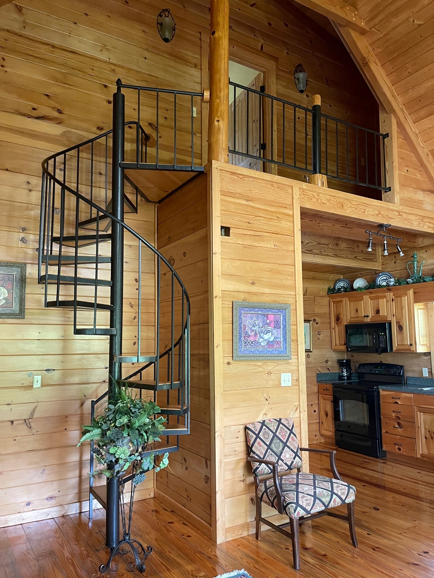 A rustic cabin interior featuring a dramatic black spiral staircase leading to a loft, exposed wood construction, and a kitchen area with wood cabinetry.