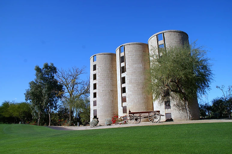 Three distinctive cylindrical stone towers with dark window openings and a wooden cart positioned in front, set against manicured green golf course turf.