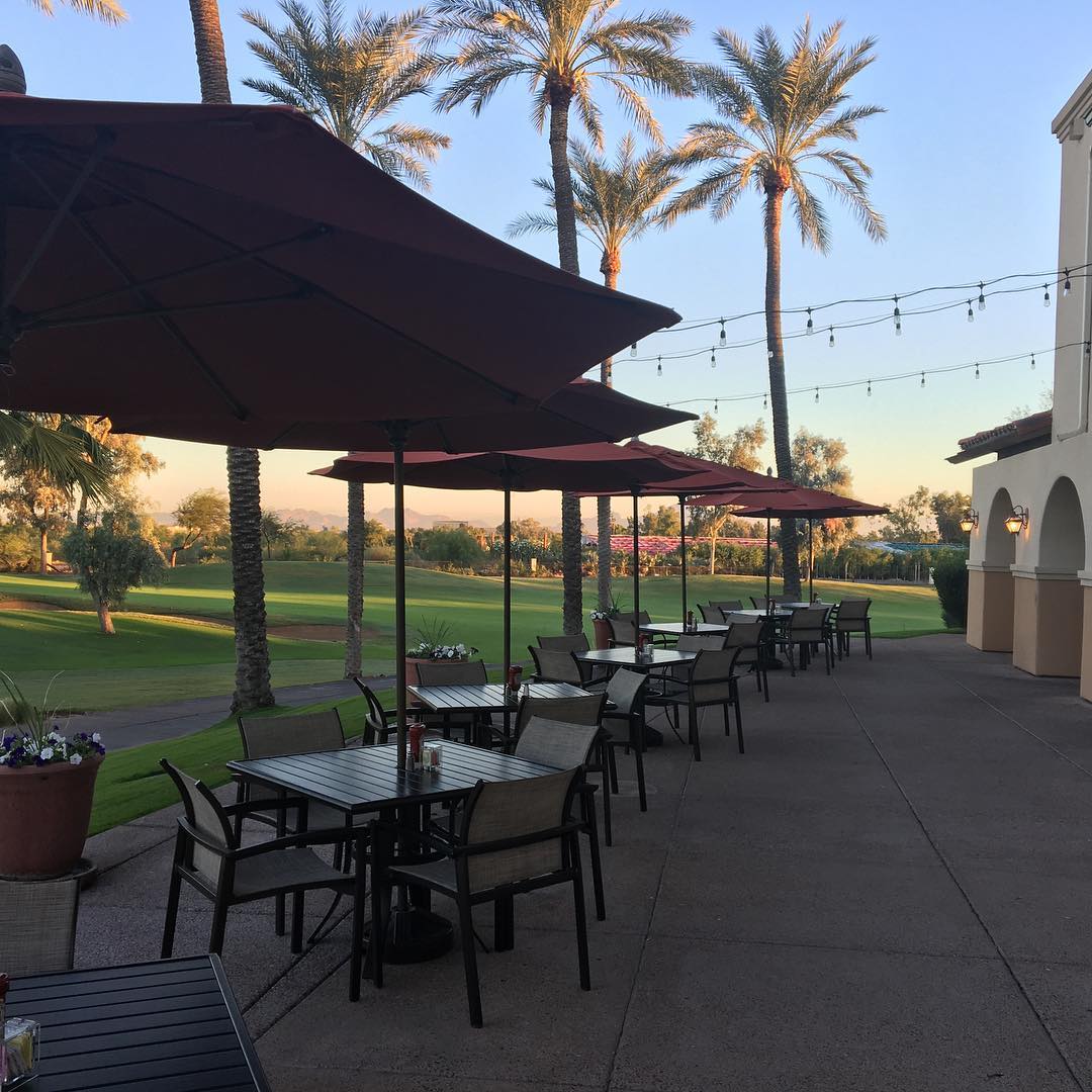 Trail's End Bar & Grill outdoor patio dining area with umbrellas overlooking the golf course during golden hour, featuring palm trees and string lights.