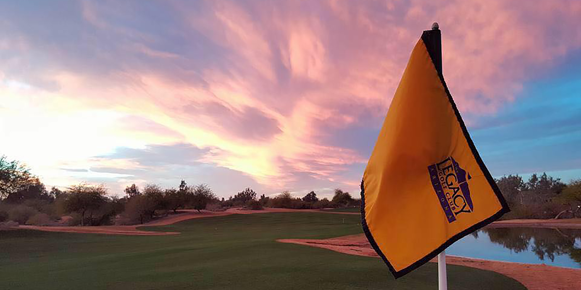 Dramatic sunset view of Legacy Golf Club with an orange Legacy flag in the foreground and golf holes visible across a water hazard with colorful sky.