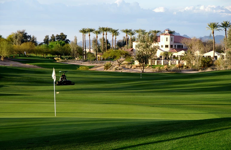 Panoramic view of Legacy Golf Resort's pristine fairway and green with the clubhouse in the background, featuring lush grass, palm trees, and desert mountains under clear skies.