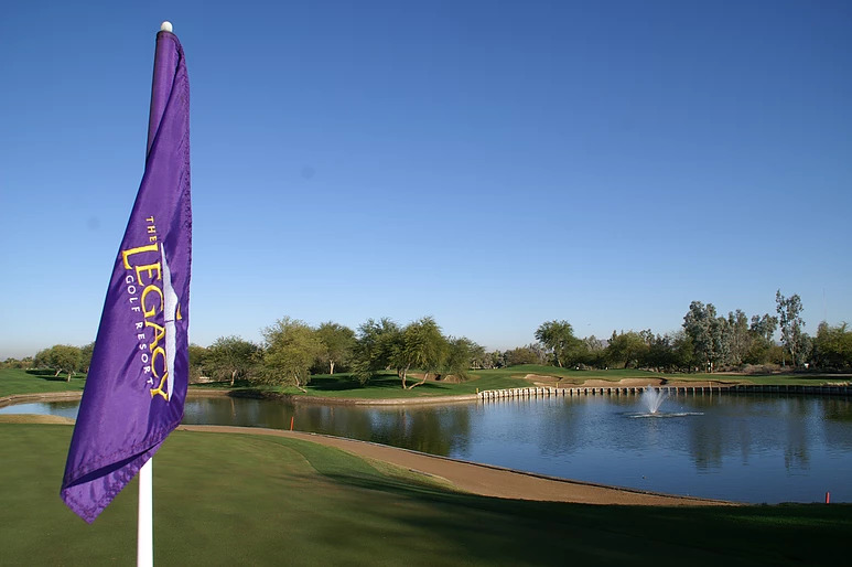 Purple Legacy Resort flag on green next to water feature with fountain, stone retaining wall, and mature trees under clear blue sky.