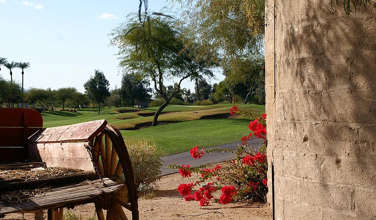 Rustic wooden cart and red flowering plants in foreground with golf course fairways and desert landscaping stretching into the background.