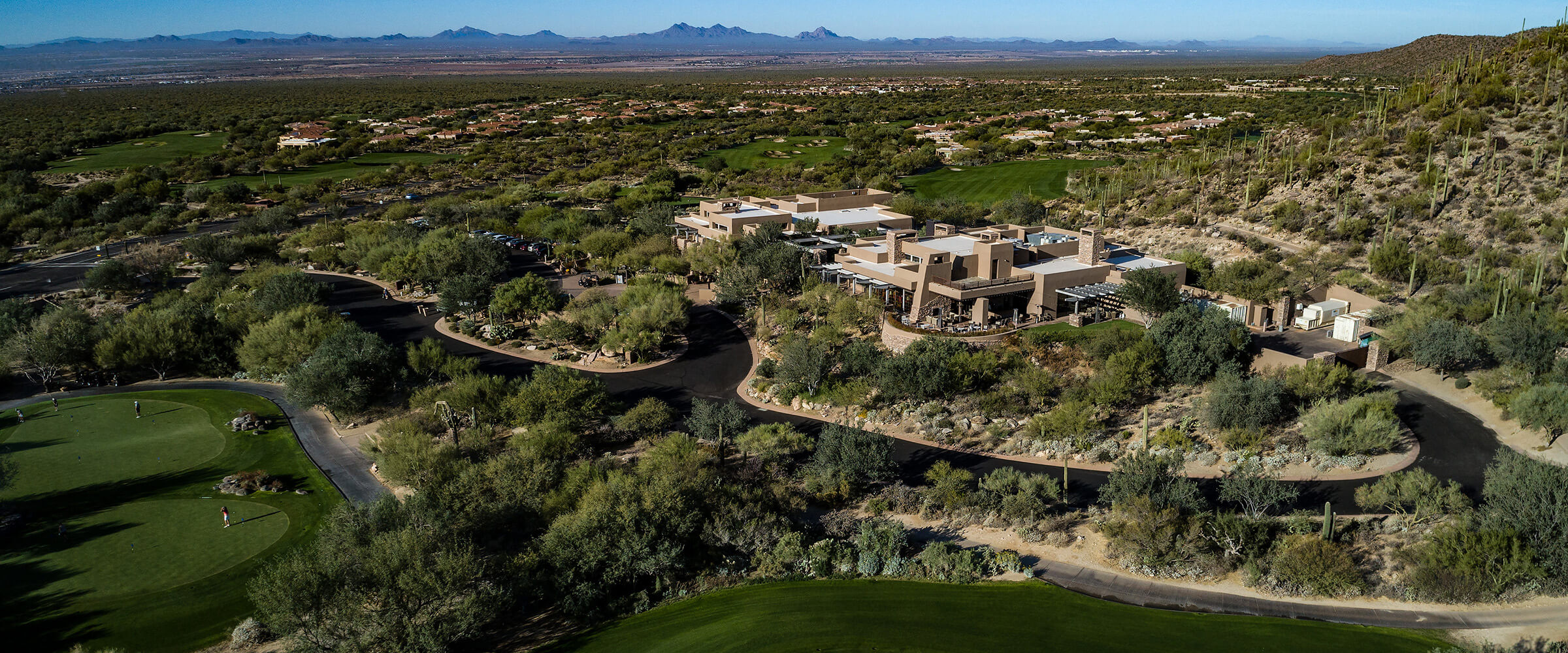 Stunning aerial view of the desert golf resort showing the modern clubhouse architecture nestled among Sonoran Desert landscape with saguaro cacti, multiple golf holes, and mountain ranges in the background.