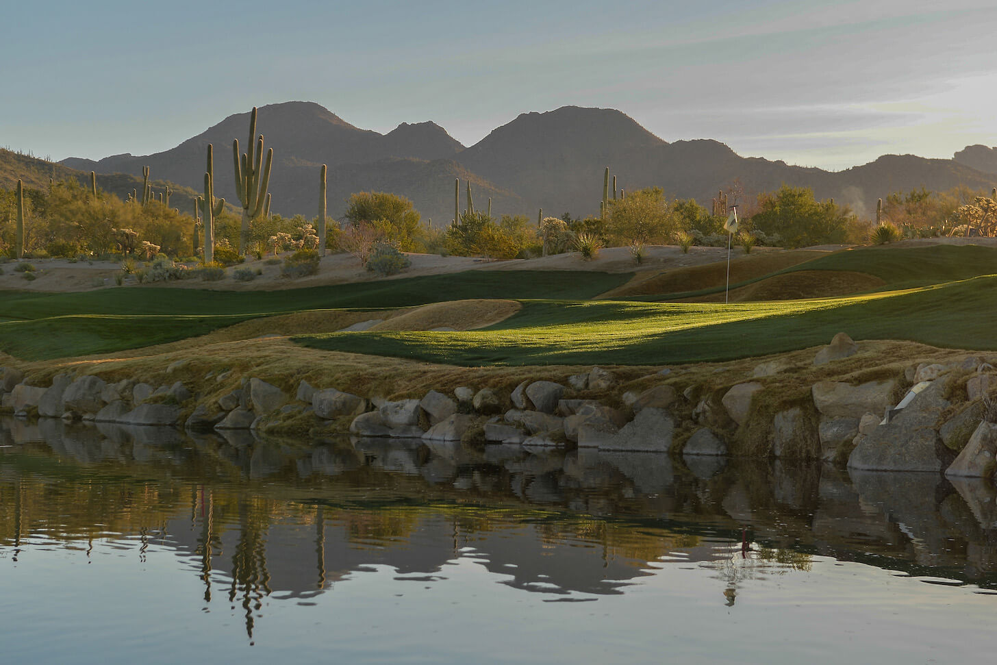 Elevated view of a challenging desert golf hole featuring strategically placed bunkers, native Sonoran Desert landscaping with multiple saguaro cacti, and expansive mountain vistas.