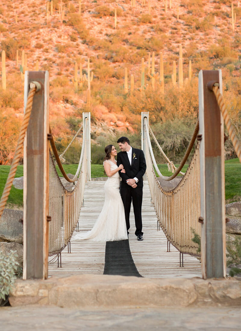 Bride and groom posing on a rustic rope suspension bridge over a desert wash, with golden-hour lighting illuminating the Sonoran Desert backdrop of saguaro cacti.