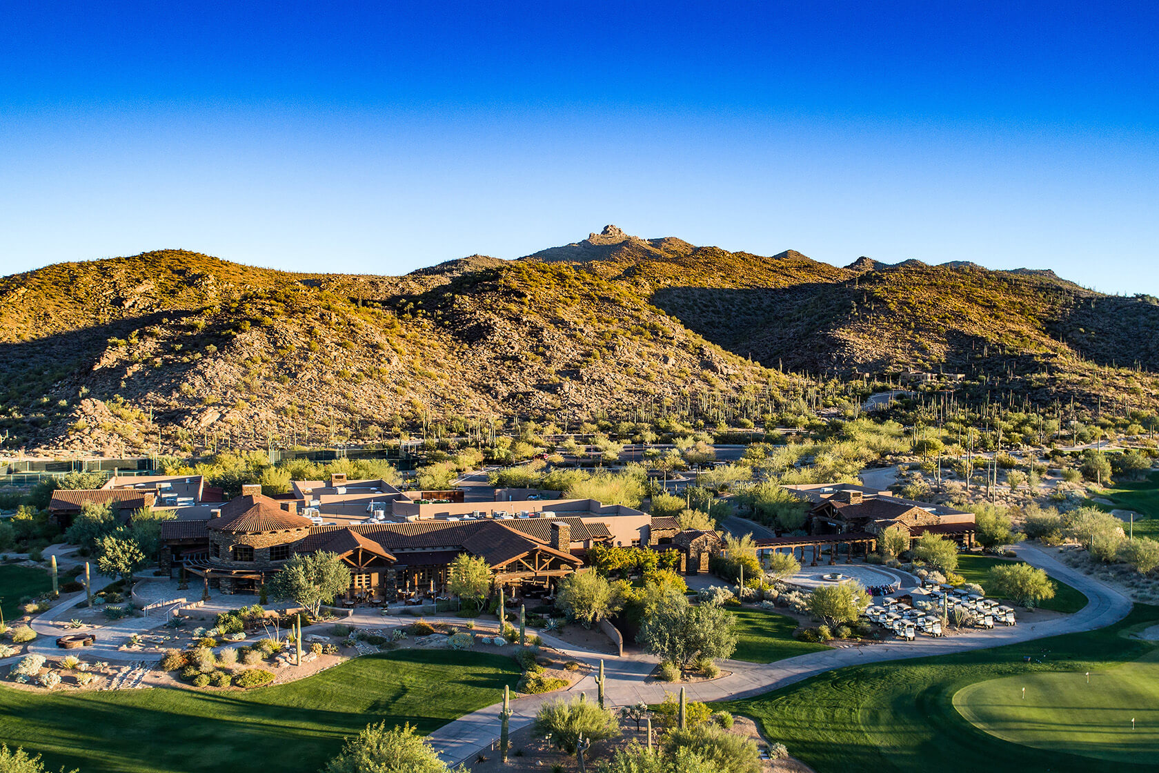 Golden hour view of the Clubs of Dove Mountain clubhouse with rustic timber architecture, golf cart staging area, and dramatic desert mountain backdrop.