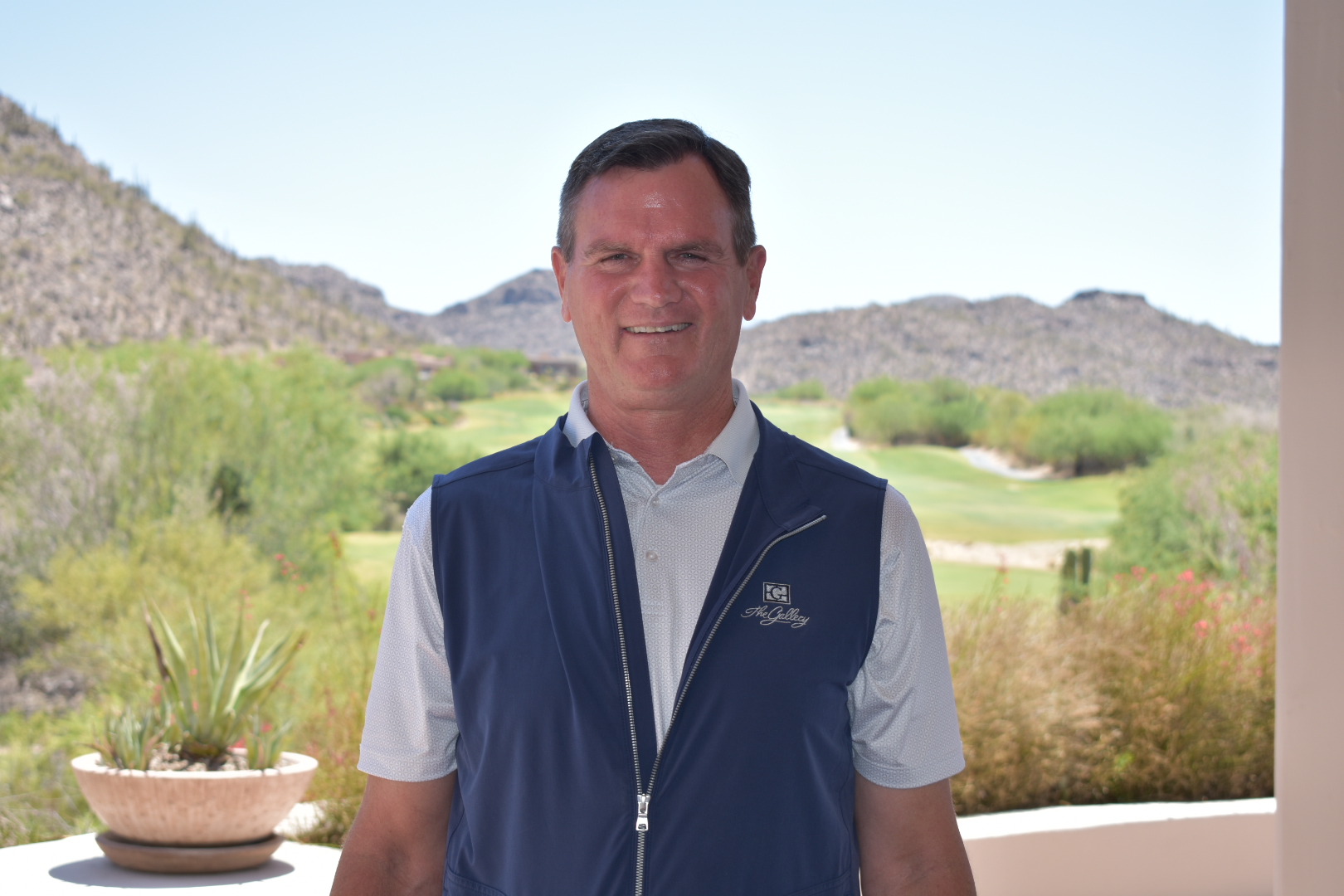 Professional headshot of a staff member in navy golf vest and white polo shirt, smiling outdoors with desert mountains and golf course visible in the background.