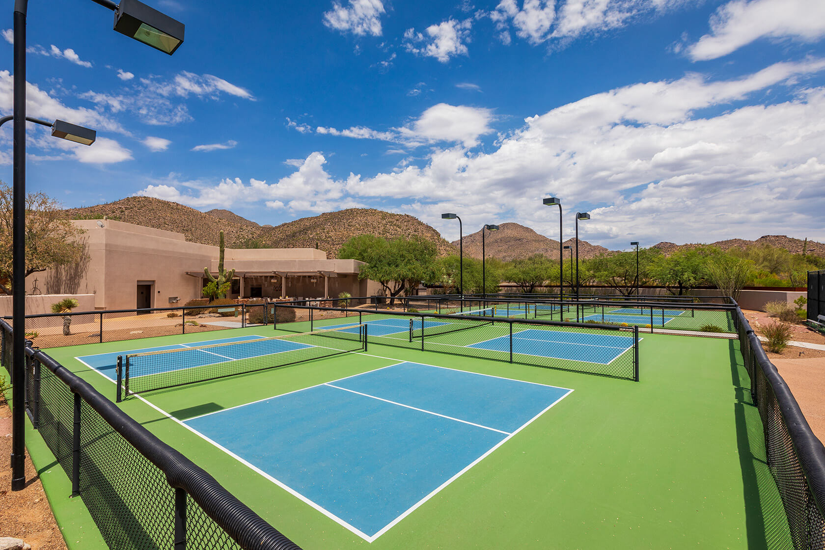 Multiple pickleball courts with bright blue and green surfaces surrounded by desert landscaping and mountain views under a dramatic cloudy sky.