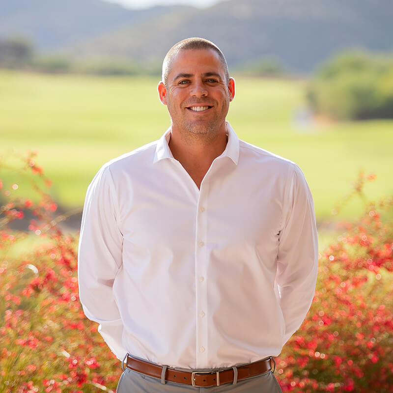 Professional headshot of Jake Hermes in crisp white dress shirt and brown belt, smiling confidently with vibrant desert wildflowers and golf course landscape in background.