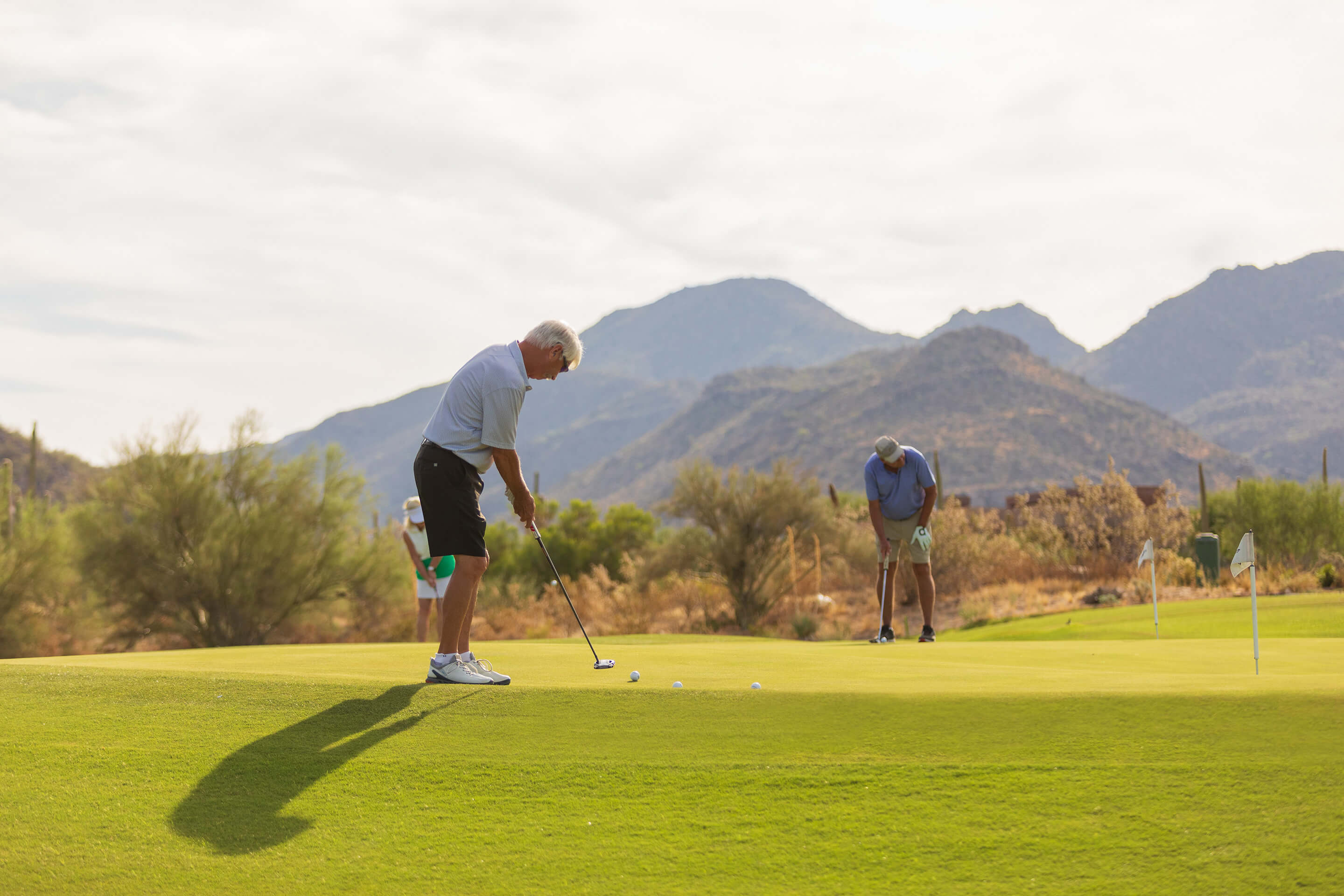 Two golfers putting on a pristine practice green with dramatic desert mountain scenery and saguaro cacti in the background during golden hour lighting.
