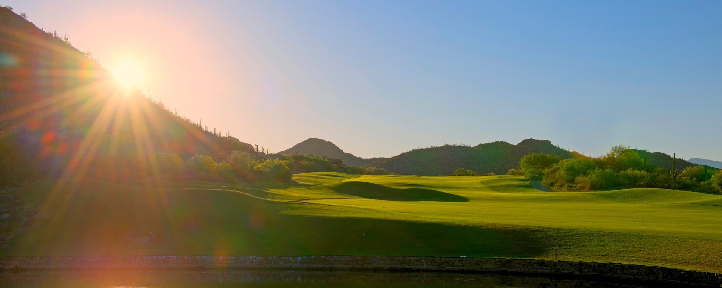 Stunning sunrise or sunset view of the golf course with dramatic lens flare, rolling fairways, and desert mountains creating a golden hour landscape.
