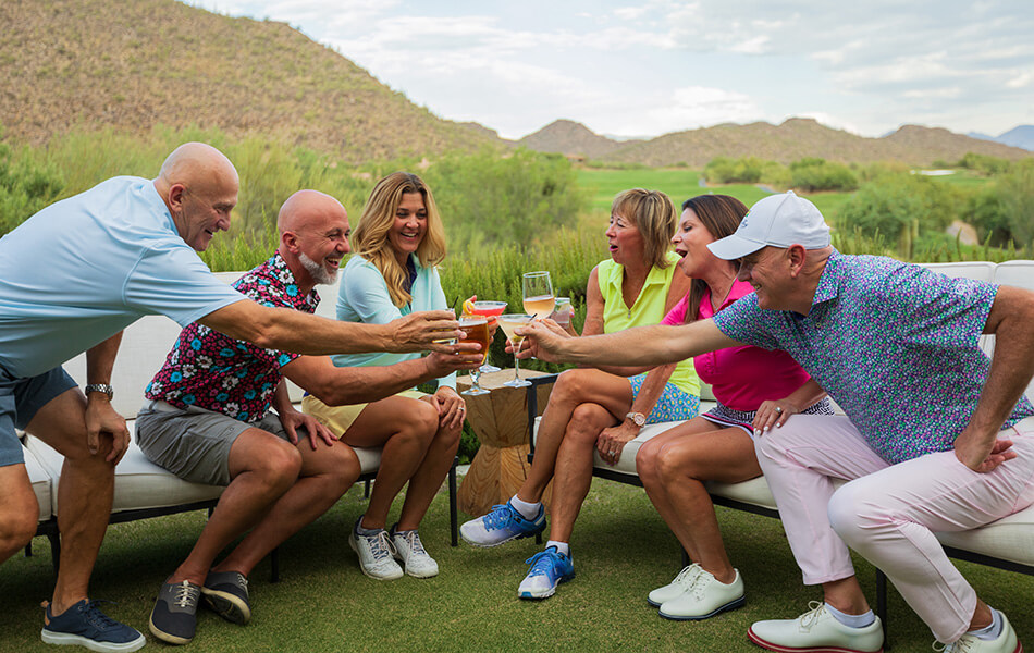 A group of club members socializing outdoors on a patio area with drinks, featuring desert mountain views and casual golf attire.