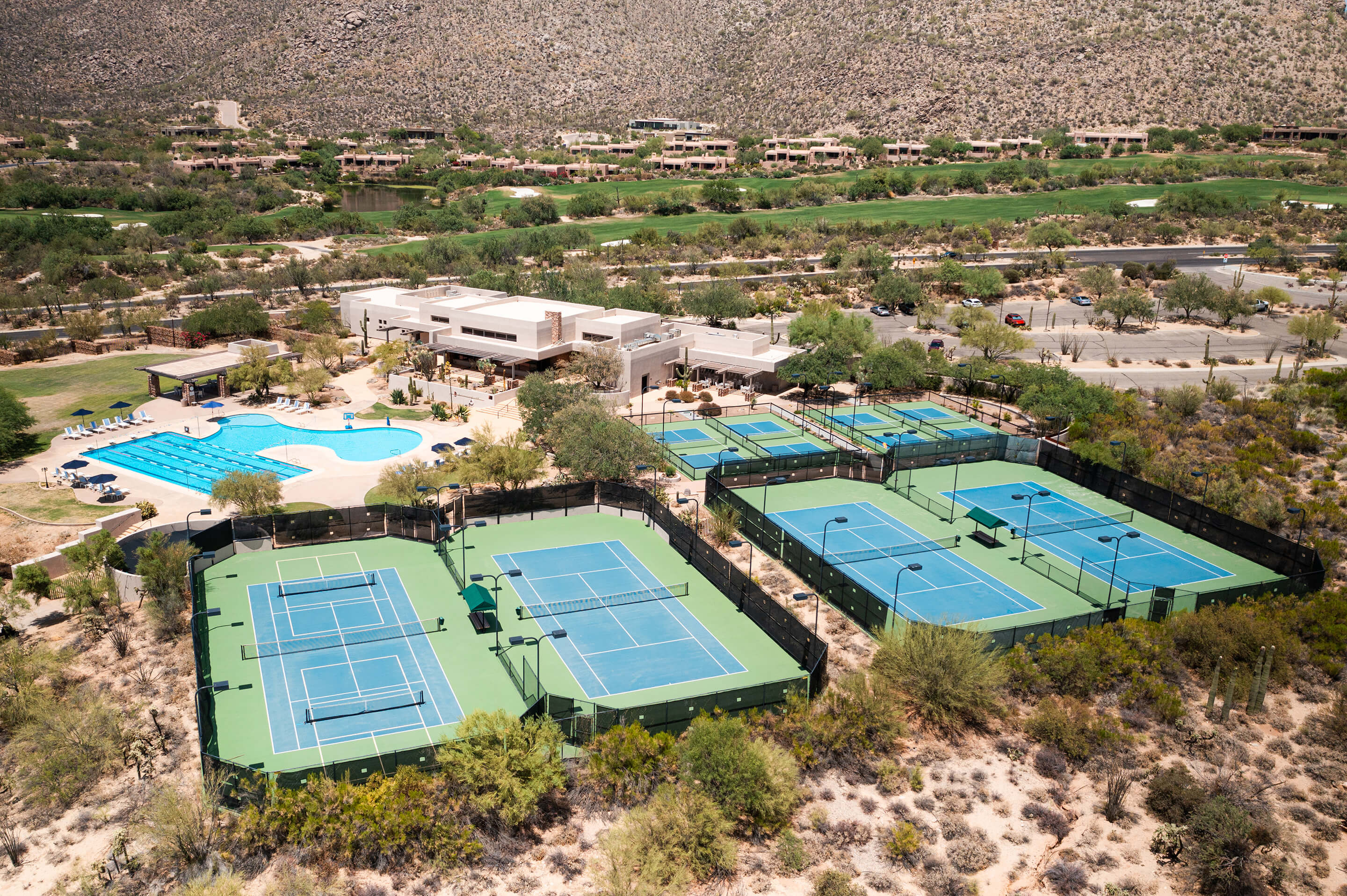 Aerial view of the club's comprehensive tennis and sports facilities including multiple courts, swimming pool, and modern clubhouse building set against desert terrain.
