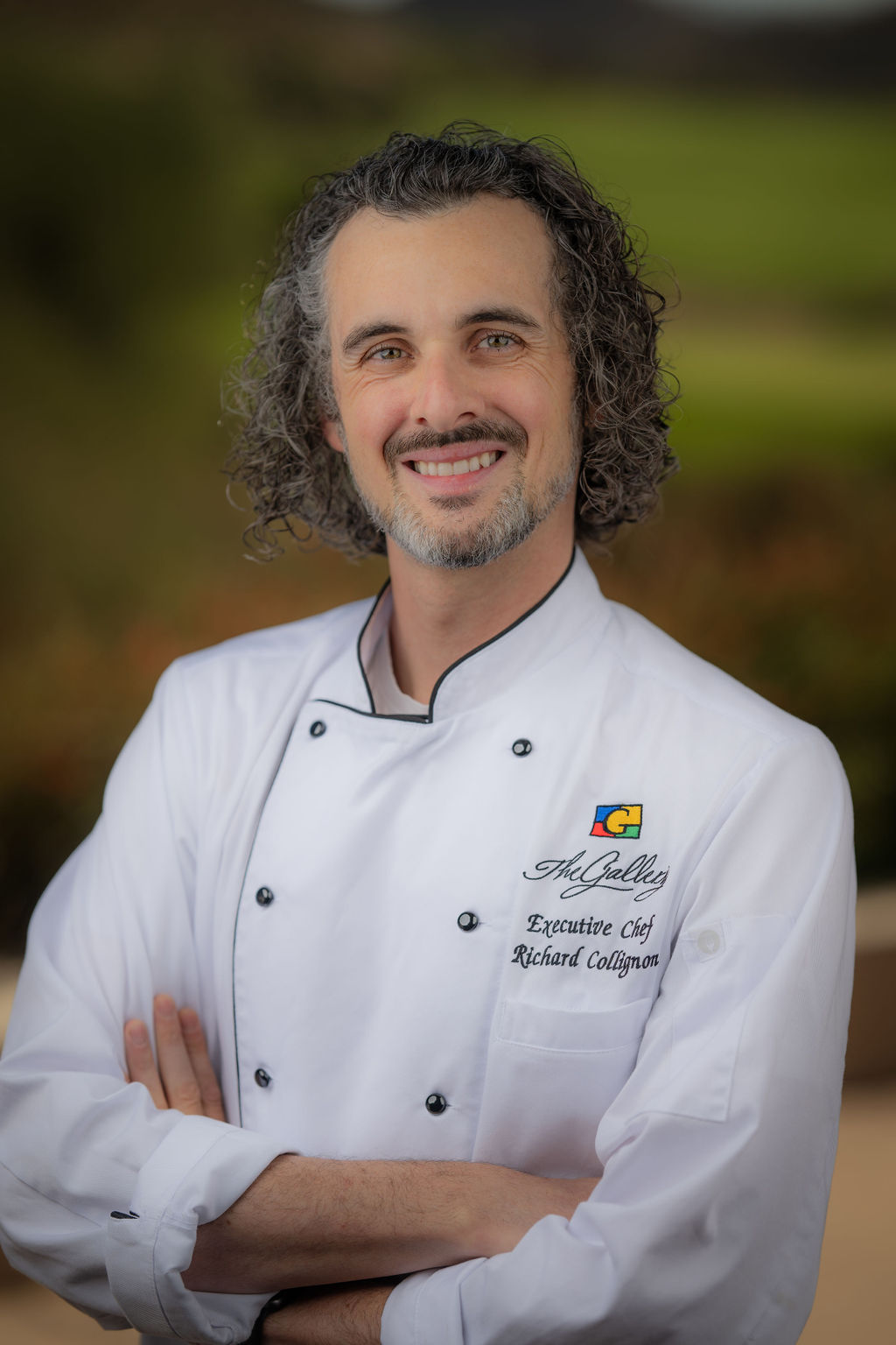 Professional headshot of Executive Chef Richard Collignon in white chef's coat with 'The Gallery' branding, smiling with arms crossed against a blurred background.