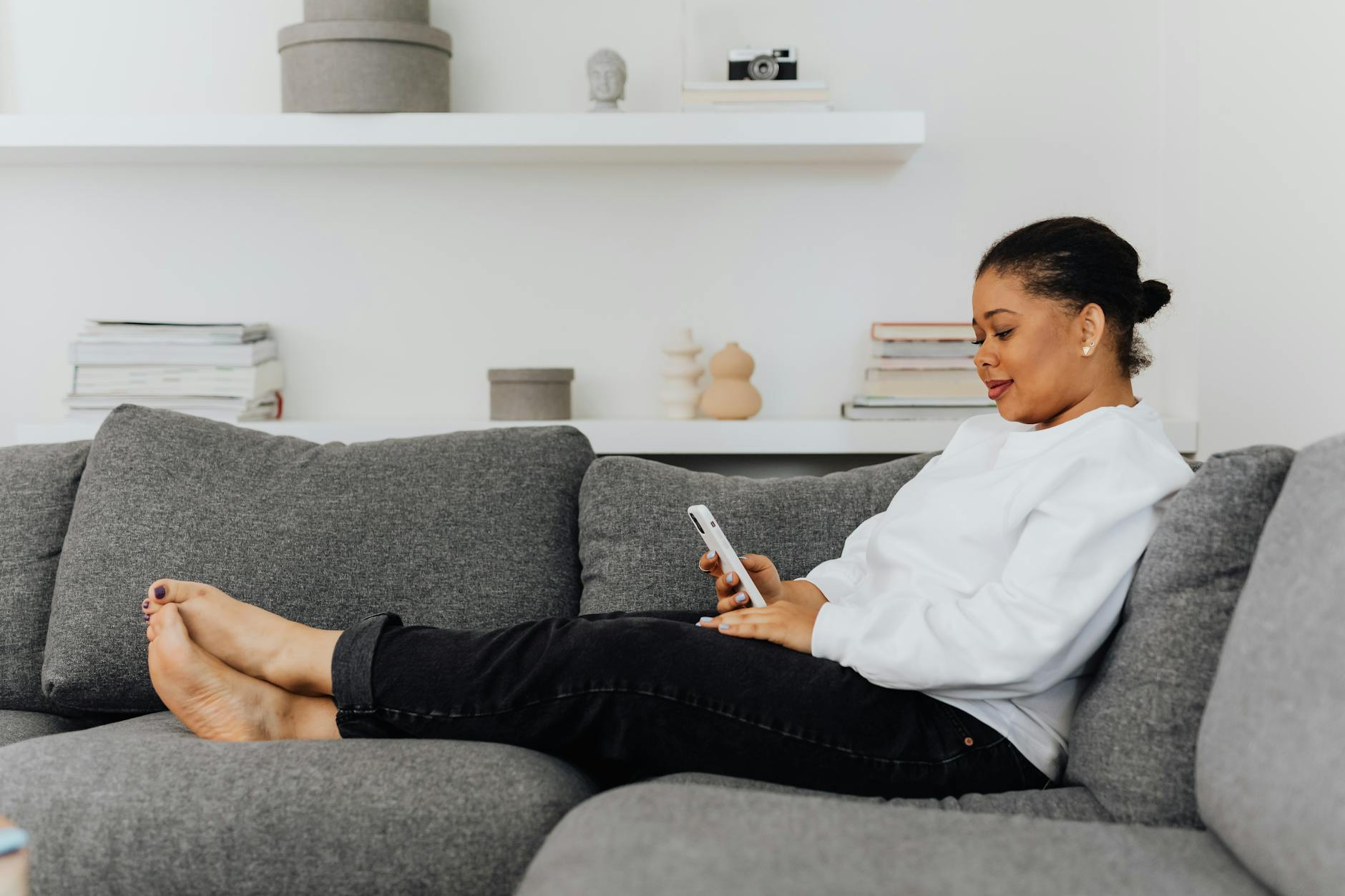 Homeowner relaxing on a gray couch browsing her phone — looking up generator questions before storm season