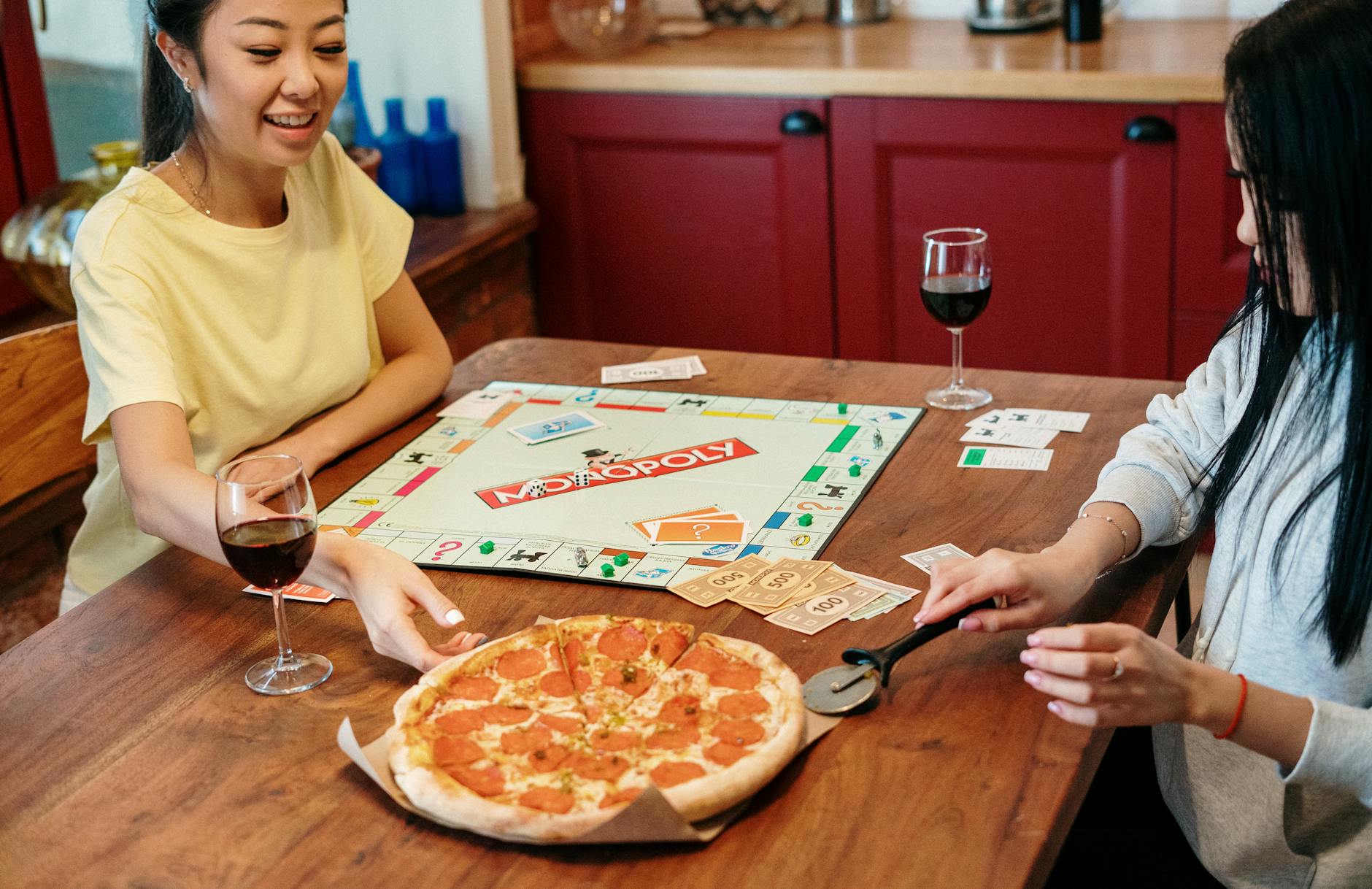 Two friends smiling over a game of Monopoly with pizza and wine on a wooden table