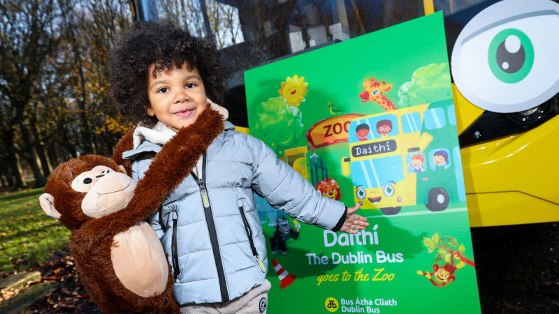 Young child with monkey backpack poses with Daithí The Dublin Bus Goes to the Zoo book poster beside yellow bus