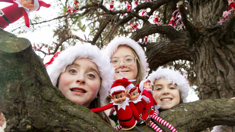 Three children in Santa hats holding elf dolls beneath a tree decorated with dozens of Christmas elf toys