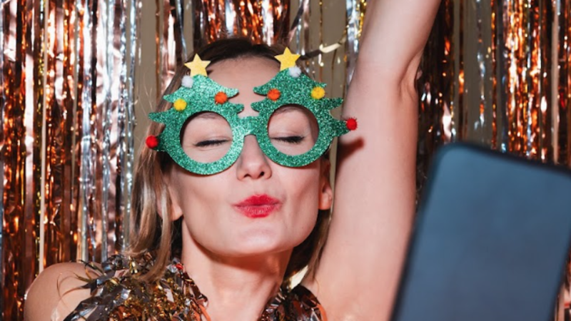 Woman wearing festive green Christmas tree novelty glasses making a playful pose at a party with tinsel backdrop