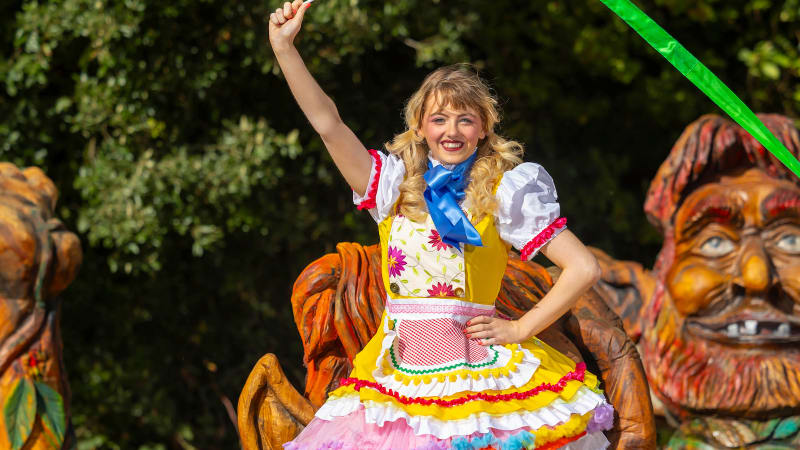 Performer in colorful dirndl costume waving green ribbon at theme park parade with decorative gnome figures