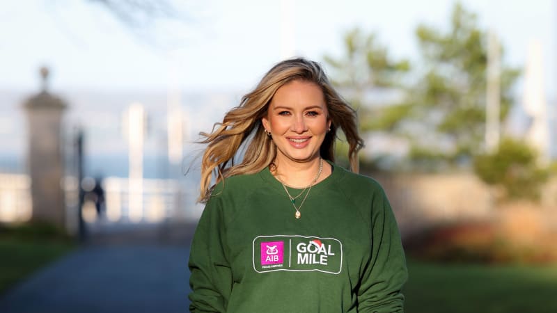 Smiling blonde woman in green AIB GOAL Mile sweatshirt outdoors on sunny day