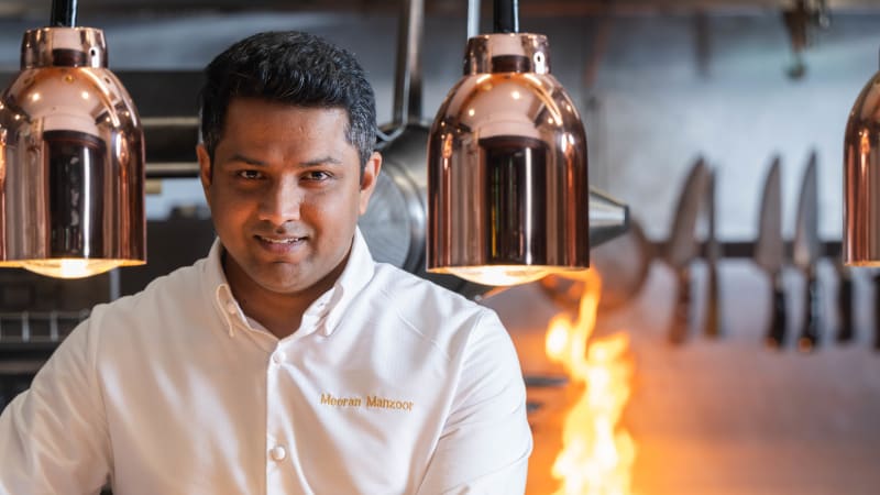 Chef Meeran Manzoor in white chef's jacket standing in professional kitchen with copper lights and flames
