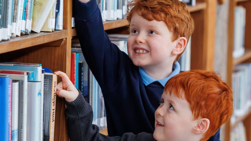 Bobby Flood and Archie Flood reaching for books on library shelves wearing school uniforms