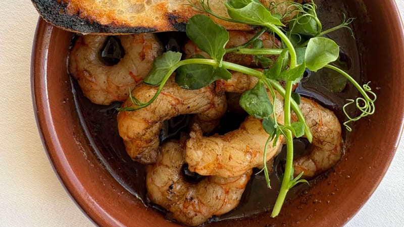 Sautéed prawns in dark sauce garnished with pea shoots, served with toasted bread in terracotta dish
