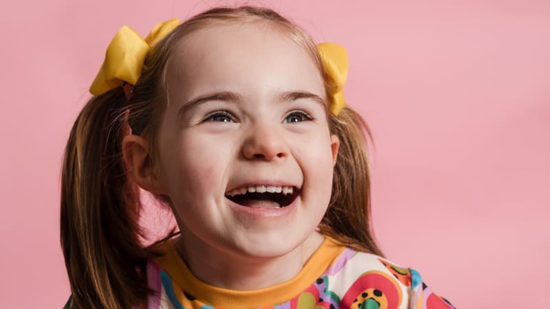 Anna, a young girl with pigtails and yellow bows, laughing against a pink background