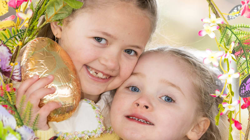 Two young blonde girls hugging and smiling, one holding a gold foil Easter egg, surrounded by floral decorations