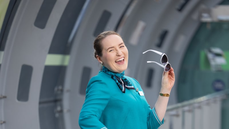 Female flight attendant in teal uniform laughing while holding white sunglasses in airport jet bridge