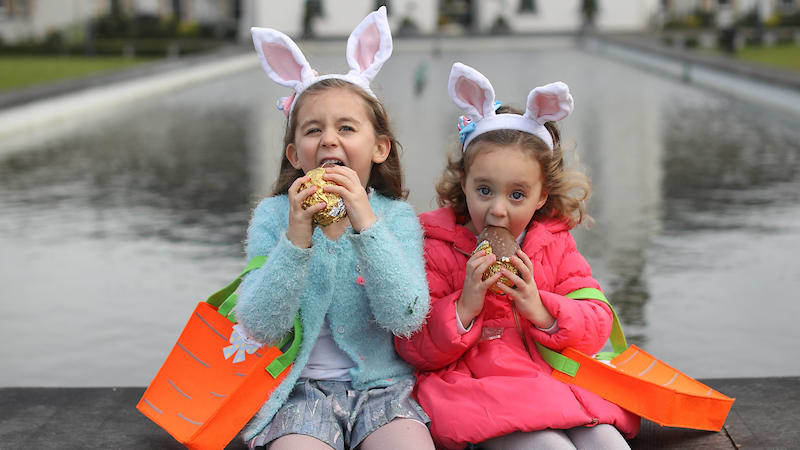 Two girls in bunny ears eating chocolate eggs with carrot-shaped Easter bags by a pool