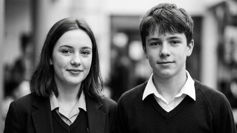 a black and white close up picture of a teenage boy and a teenage girls side by side looking at the camera. relaxed looking. Just shoulder to shoulder from the shoulders up in shot. no arms around each other. wearing school uniform