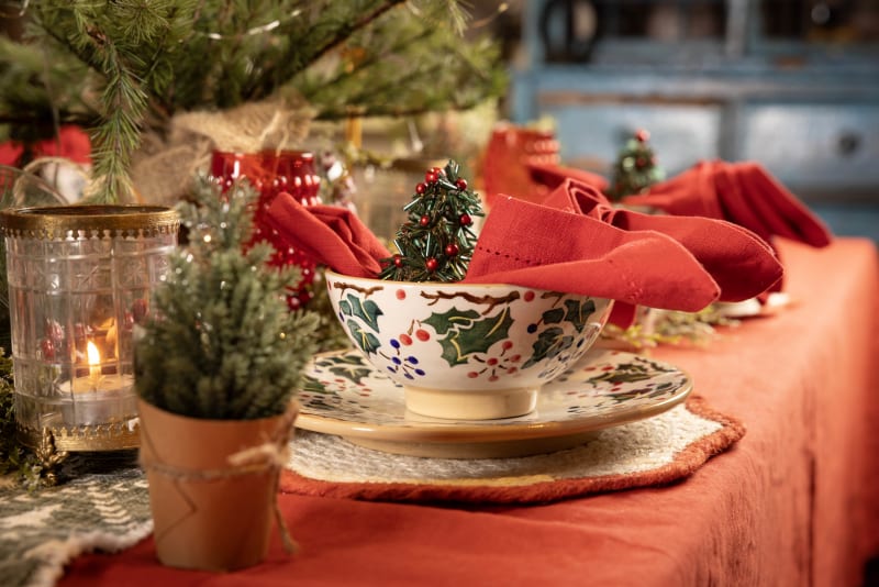 Christmas table setting with holly-patterned bowl and plates, red napkin, candle, and festive greenery decorations