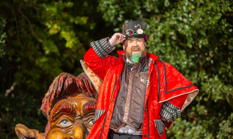 Man in steampunk costume with green beard and red cape posing next to carved wooden goblin sculpture