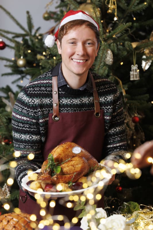 Young man in Santa hat and Christmas sweater smiling in front of decorated tree