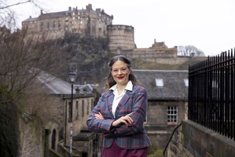 Debi Fry standing with arms crossed in tartan blazer with Edinburgh Castle in background