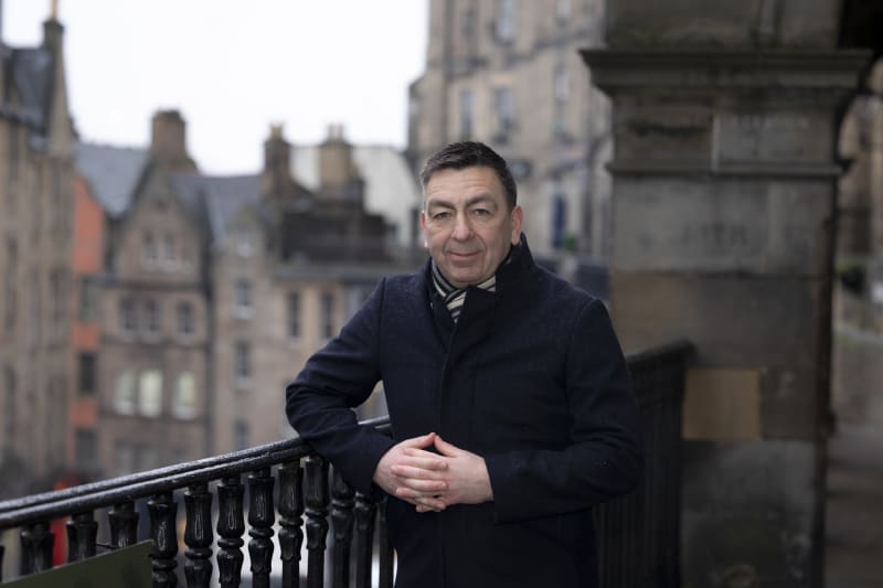 Paul Stanfield in a navy coat standing on a balcony with Edinburgh's historic buildings behind him